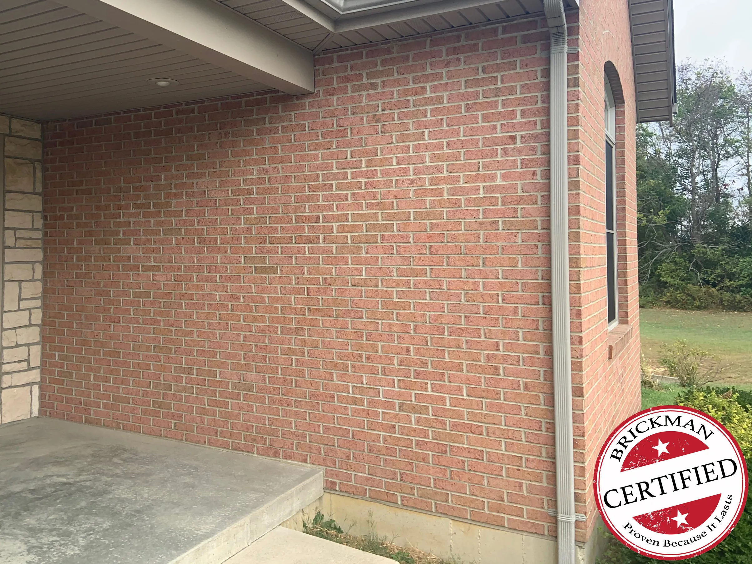 The same covered entrance wall in 2019, one even range of pink and tan brick running to the stone door surround, with the certification stamp in the corner