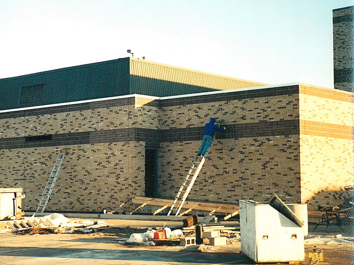 An applicator on a ladder working along the dark band at the corner of the school in 2001, with ladders, timber and a site bin on the tarmac below