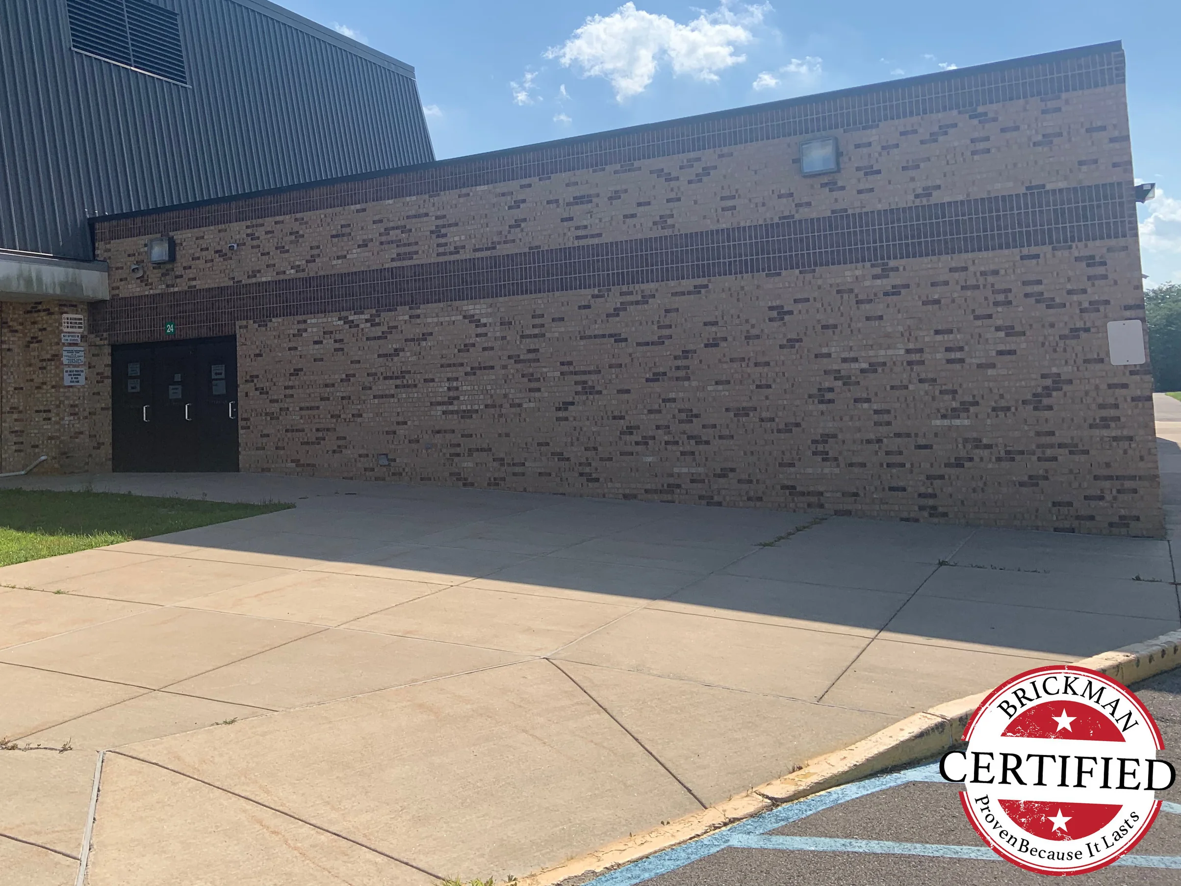 A low brick wing of the school in 2019 with a tall gymnasium block behind it, one dark band running the length of the buff brick wall and the Brickman certified stamp in the corner of the frame