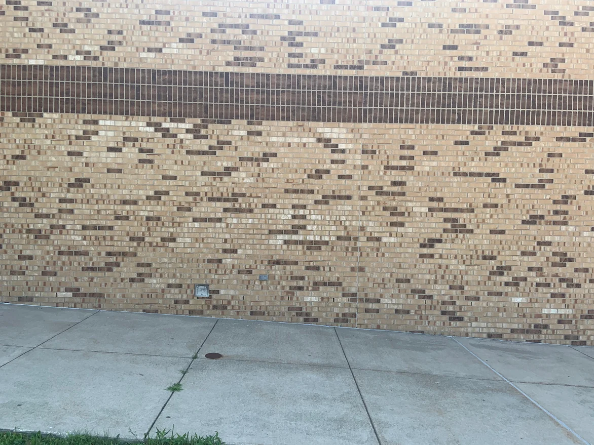 A stretch of the school wall in 2019 with the dark soldier band across the top, dark headers scattered through the buff brick and a concrete walk along its base