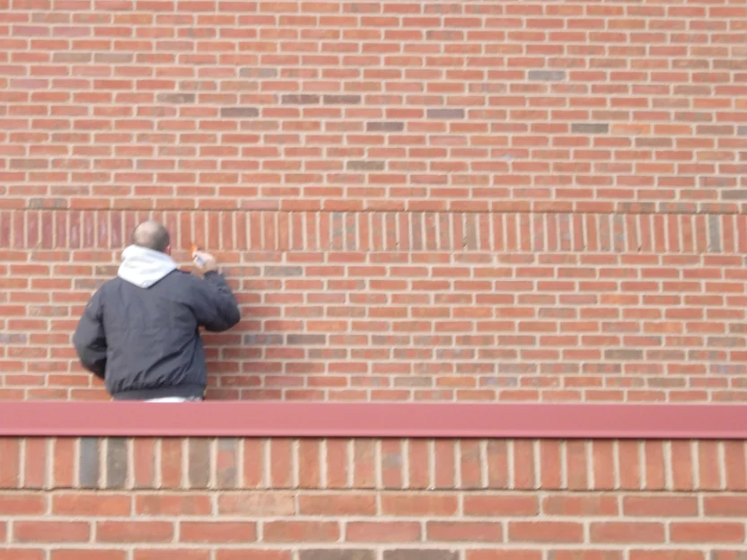 An applicator working by hand along a course of upright brick on a large red brick wall, standing at a low parapet