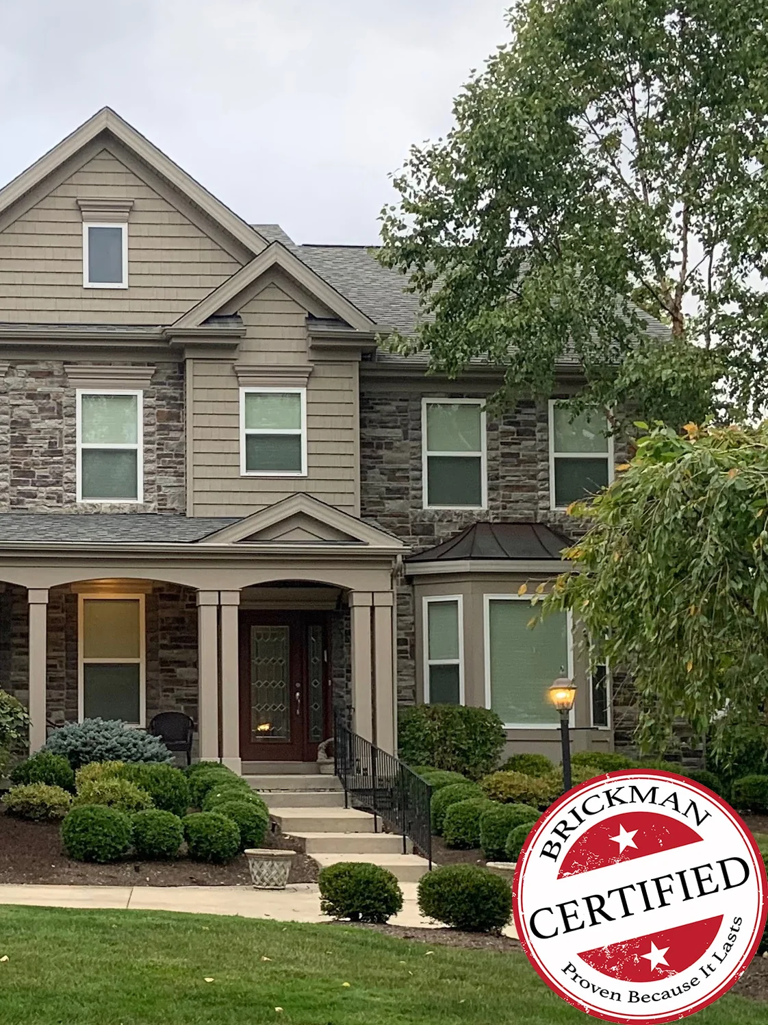 The front of the house in 2020, the window sills now a grey that sits inside the stone blend, with mature planting below and the certification stamp in the corner