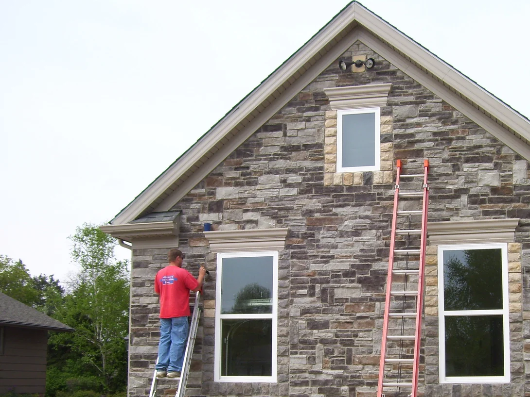 The gable end of the house in 2009, an applicator on a ladder working stain into the tan stone beside a window while a second ladder stands against the wall