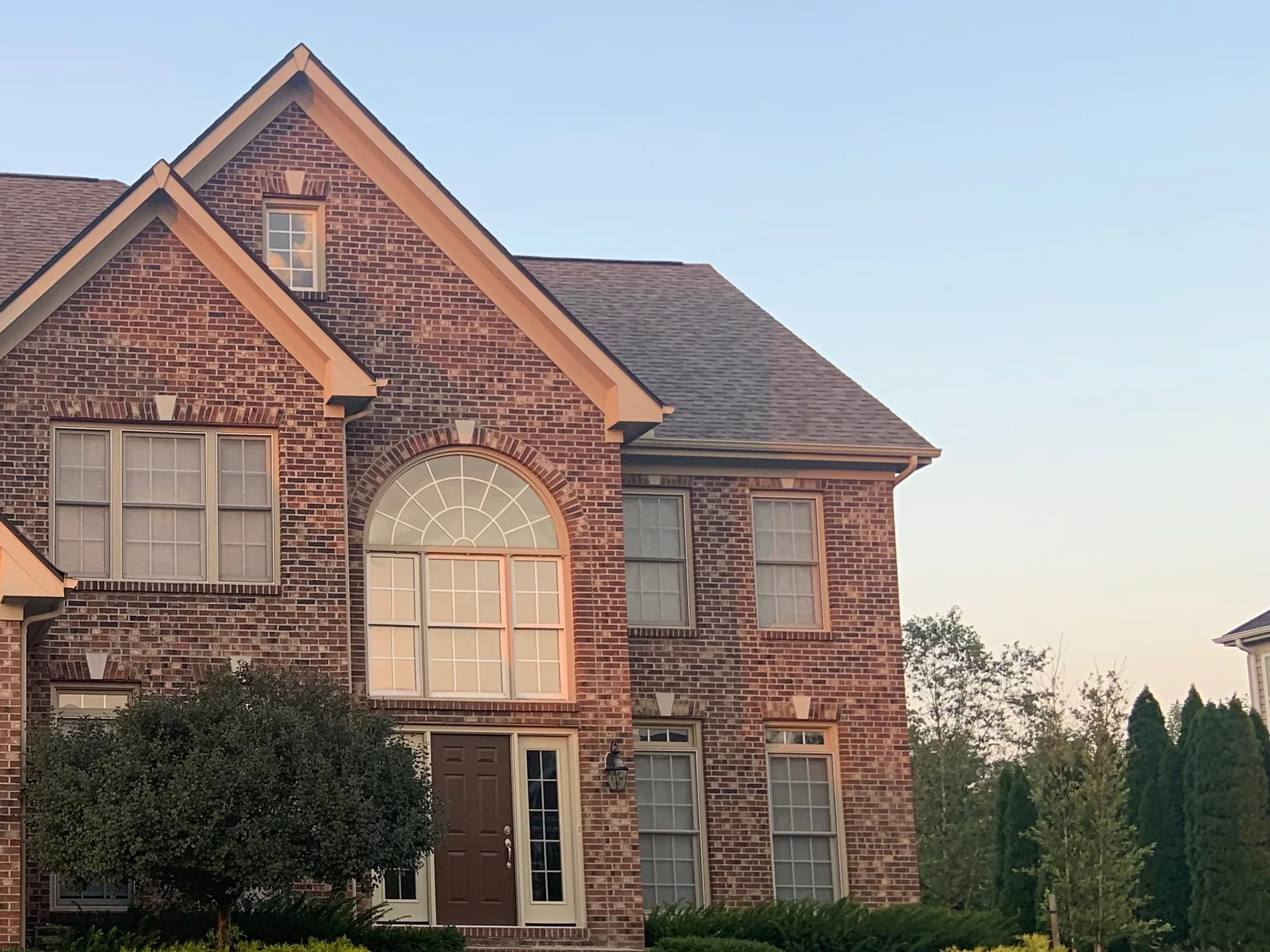 The front of the house in 2019, an arched entry window under a tall gable and a two-story wing beside it, the brick reading as an even range of reds and browns from the eaves down to the planting