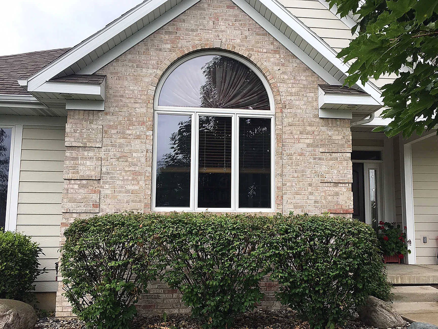 The front gable of the house in 2019, its brick reading as one even blend from the arched window down to the shrub bed