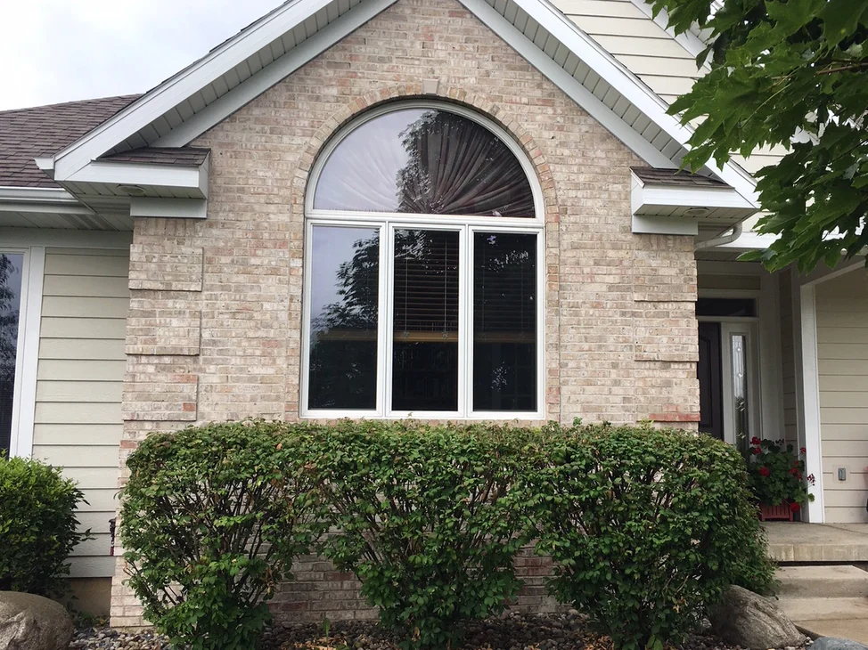 The front gable of the house in 2019, its brick reading as one even blend from the arched window down to the shrub bed.