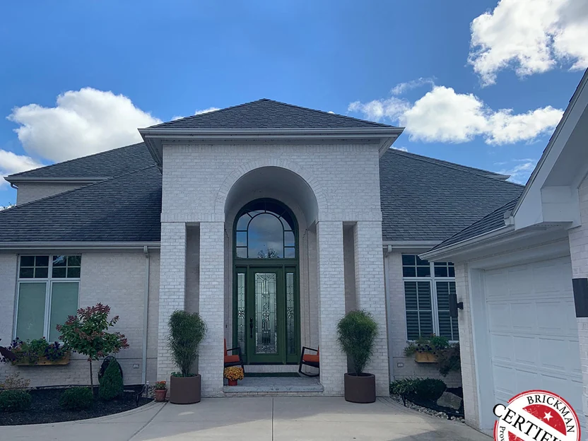 The front of the house in 2019, white brick under a dark hip roof, a green front door inside the arched portico with planters either side, the certification stamp in the corner