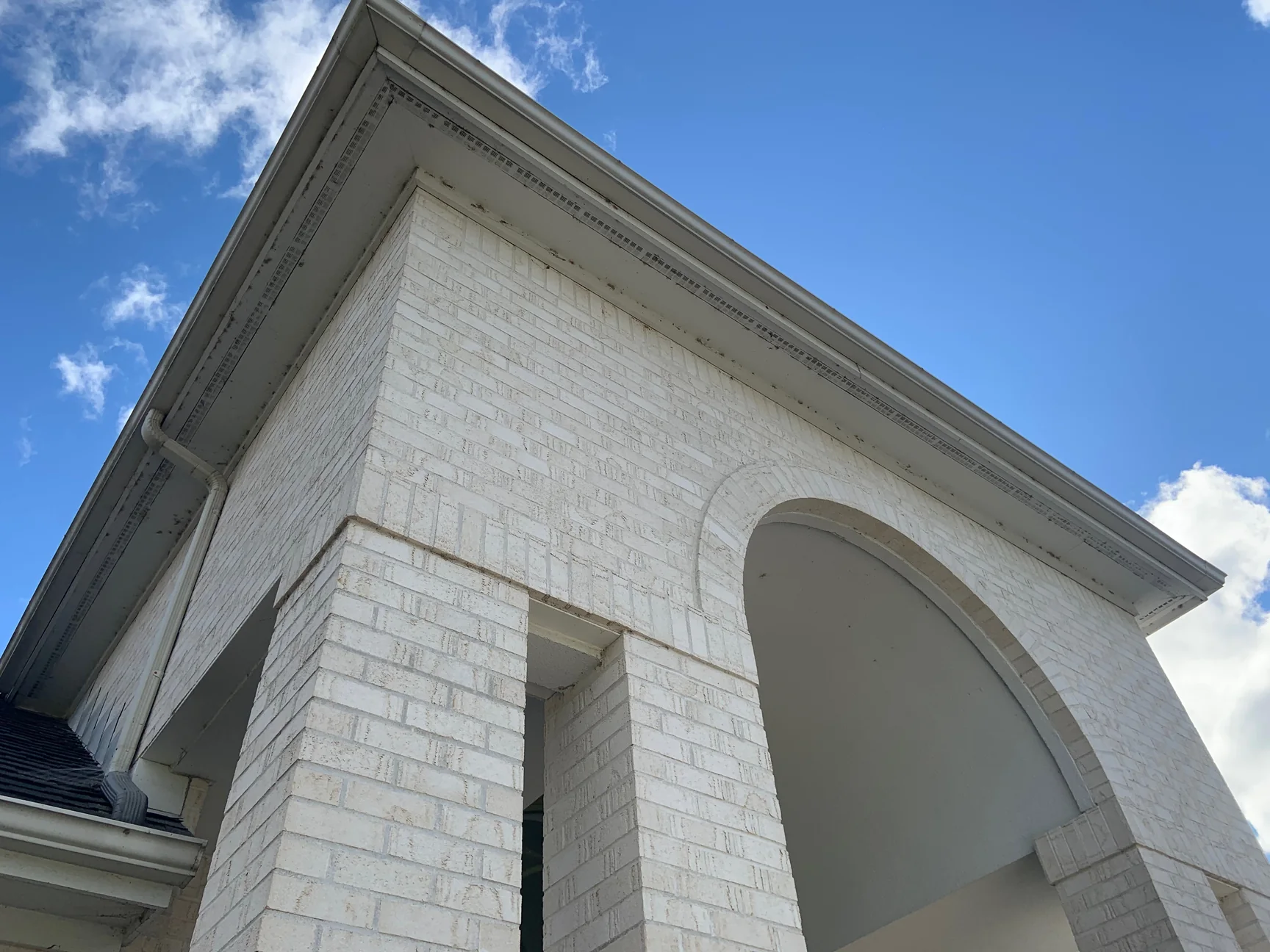 The entry portico in 2019 looking up from the driveway, white brick piers and an arch under a dentil cornice and gutter, blue sky and cloud above
