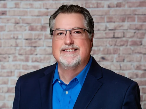 Studio headshot of Don Foster in a navy blazer against a light brick wall