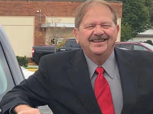 Rick Conner in a dark suit, leaning on a pickup truck in a parking lot