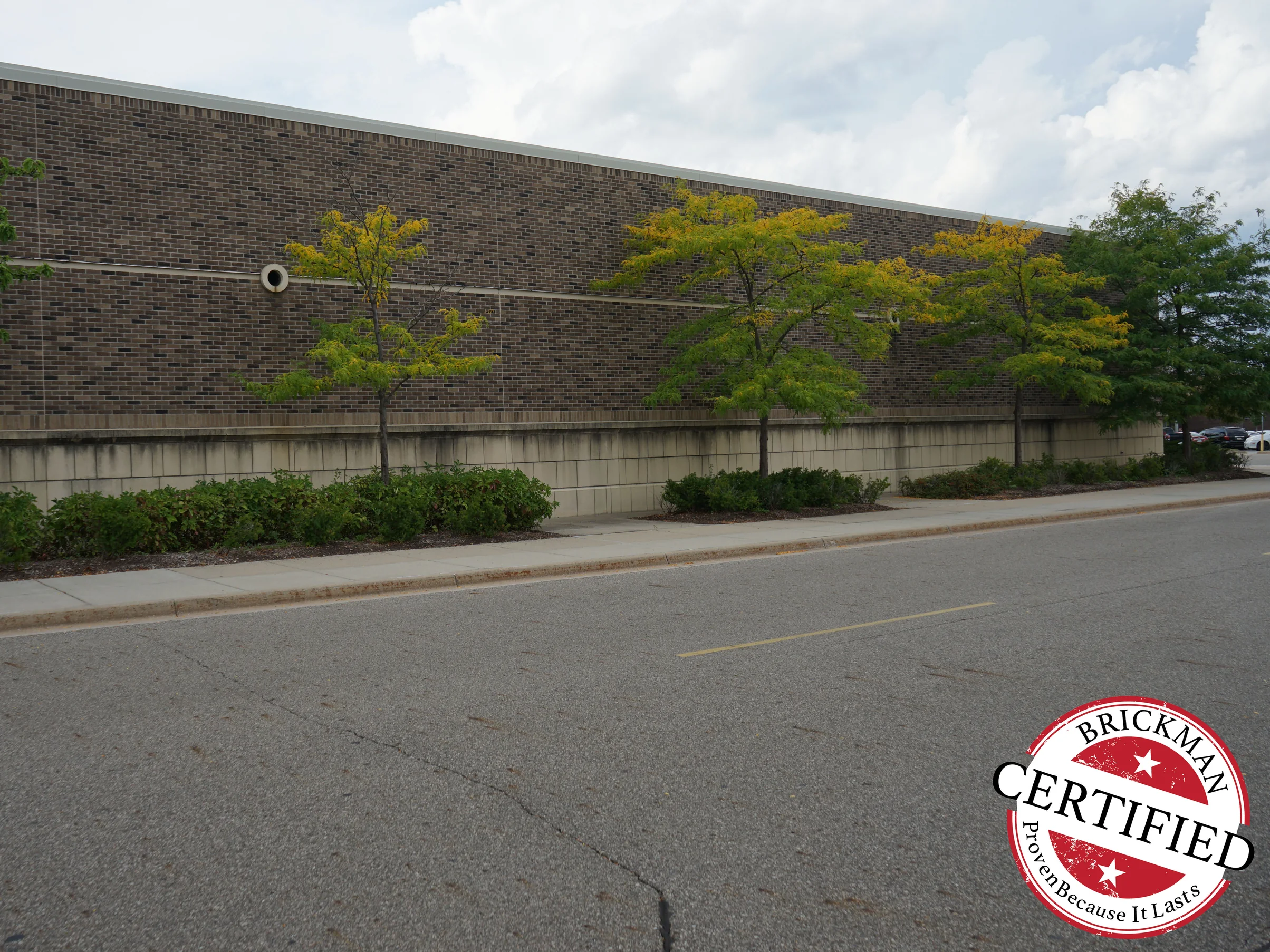 The mall wall looked along from the service drive in 2019, the brick holding one brown and tan range for the full run of the elevation behind a row of young trees, the frame carrying the Brickman Certified stamp
