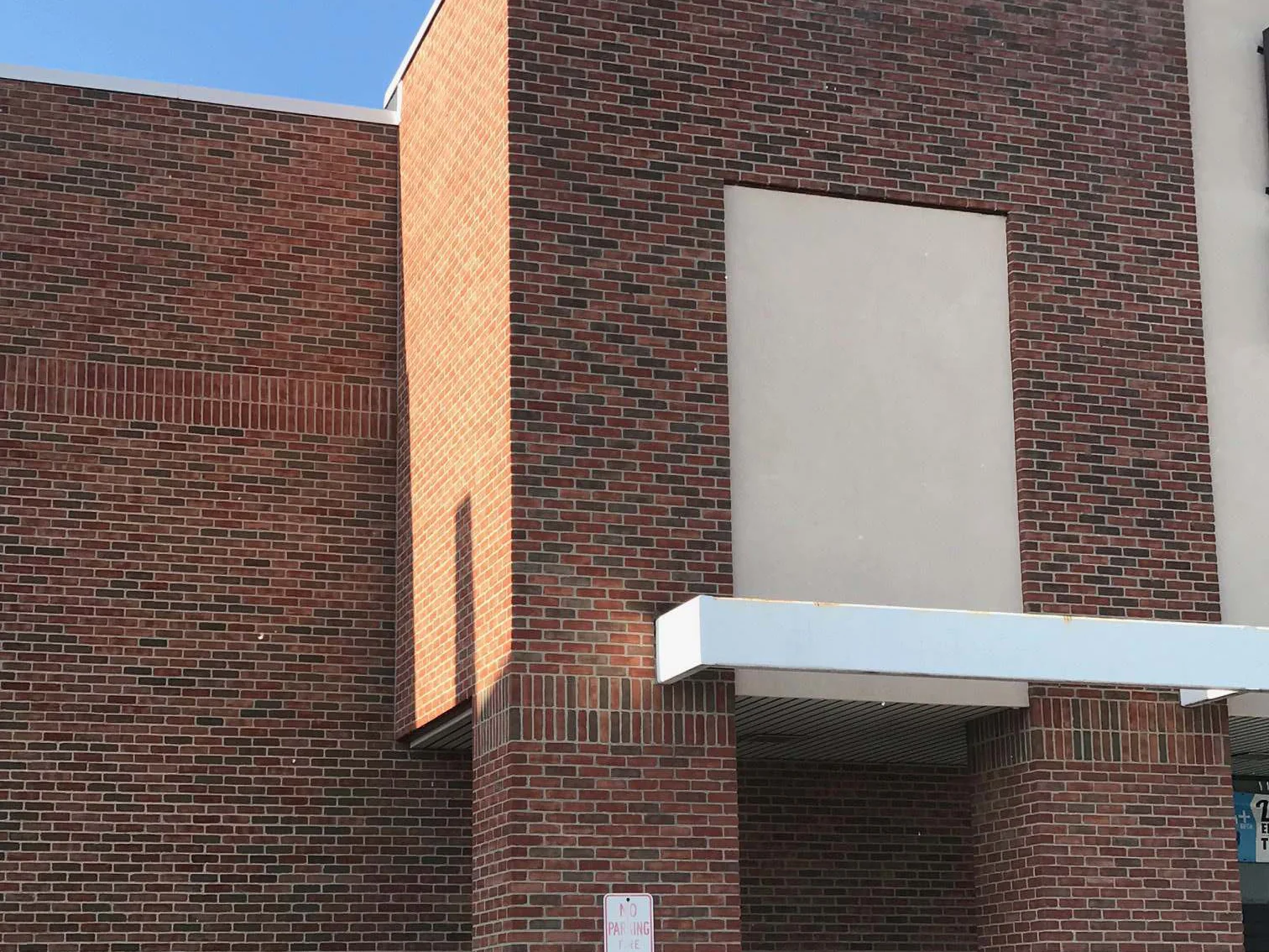 The entrance tower and the wall beside it in 2019, one flashed red brown blend across both, with a blank stucco panel, a white canopy and a no parking sign on the pier