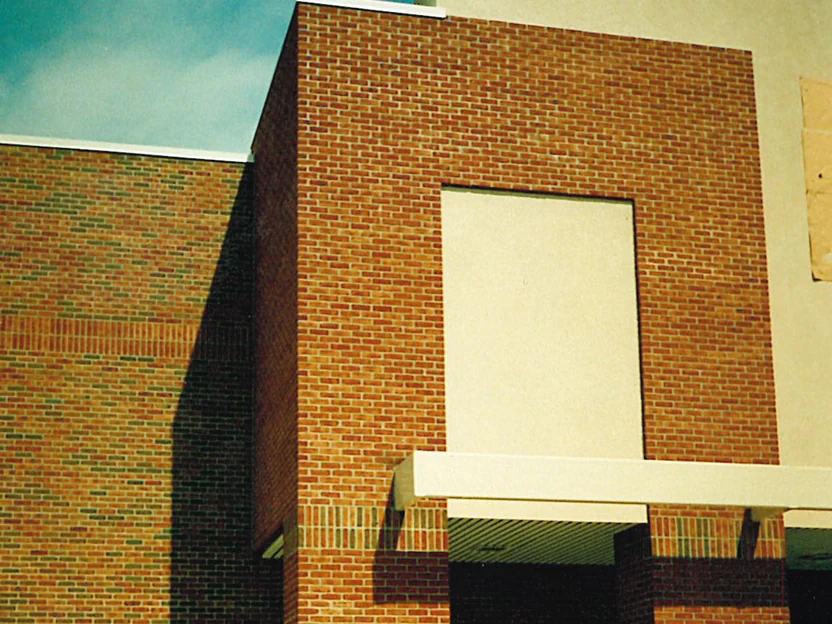 The entrance tower in 1995 before staining, plain orange tan brick around a blank stucco panel and a white canopy, beside a wall of darker flashed brick to the left