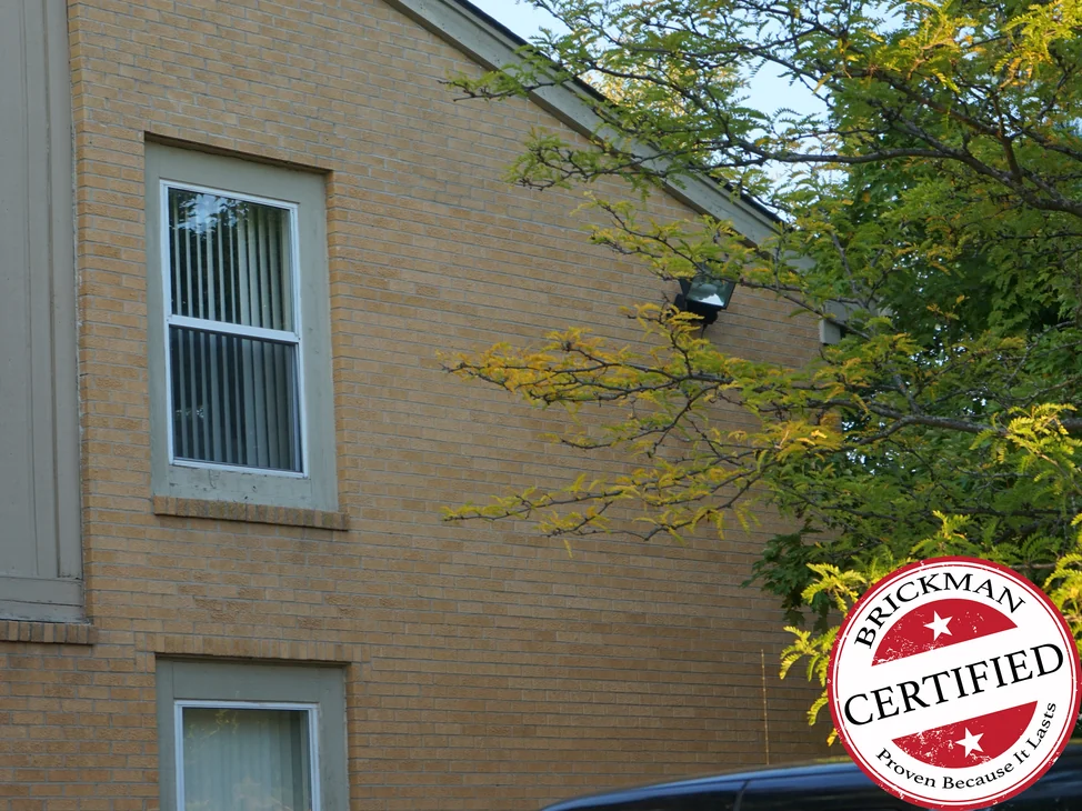 The buff brick gable end of a Michigan apartment block in 2019, one even range from the ground to the raking eaves with the Brickman Certified stamp at the lower right.