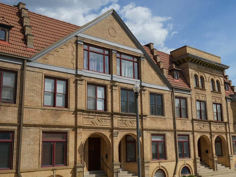 The street elevation in 2019, buff brick running past a gabled bay and a run of arched entries under a red tile roof