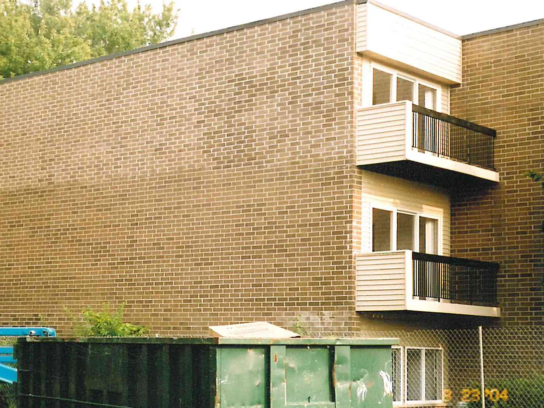 The blank end wall of a three-story apartment block in 2004, balconies and windows stepping down its right-hand corner above a fenced enclosure