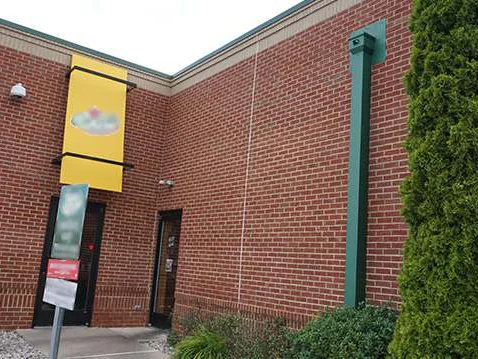 The service door wall in 2019, a yellow banner over the entrance and an evergreen grown up against the corner, the courses one red range down to the buff base