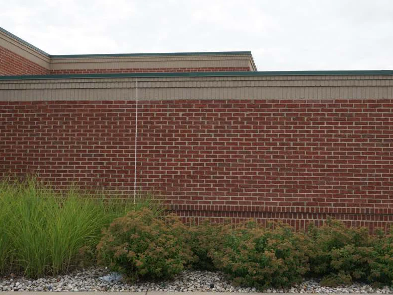 A long side wall in 2019, red brick running even from the buff band under the roofline to the shrubs and grasses at its base
