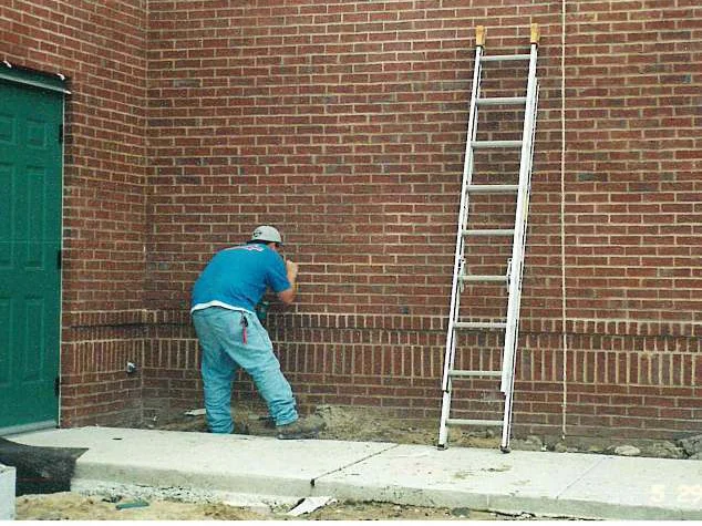 An applicator working stain into the brick by hand at the base of a wall in 2001, an extension ladder set against the courses beside him