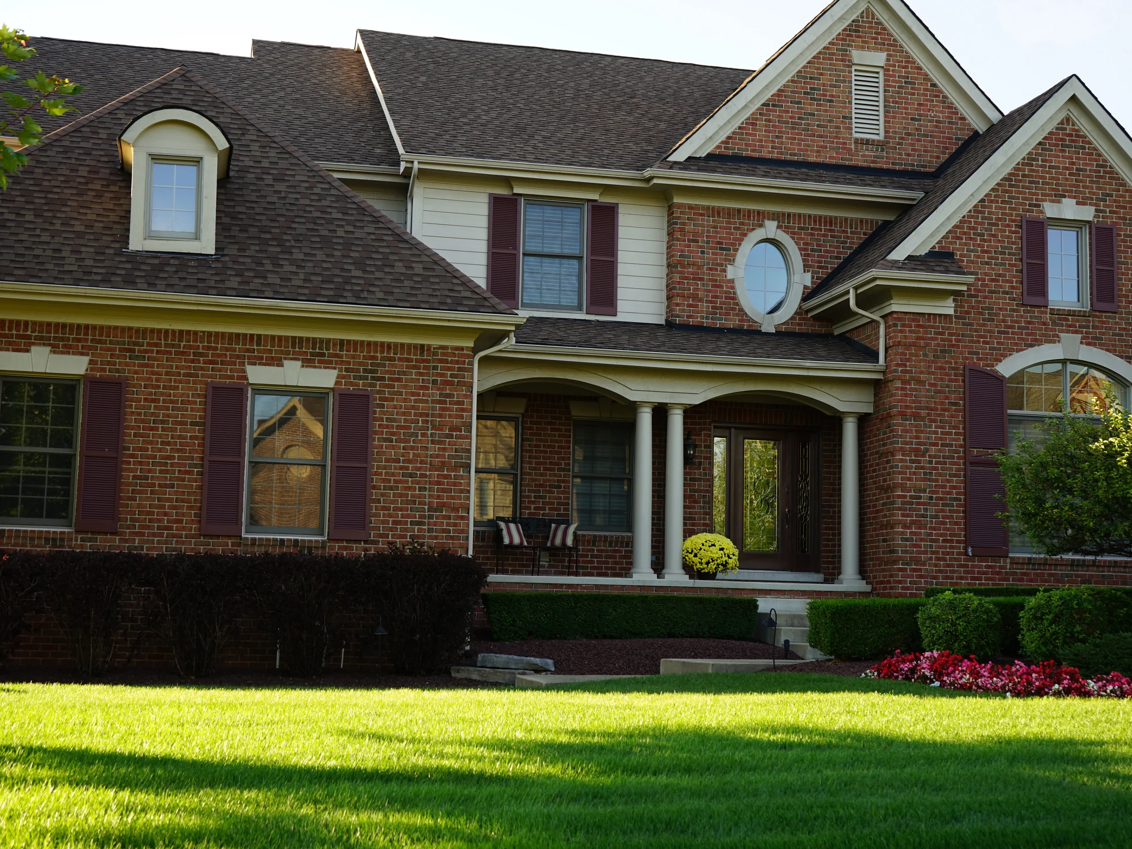 The front of the house in 2019 across the lawn, brick with cast stone lintels, an oval window over the columned porch, the wall reading as one red and brown range