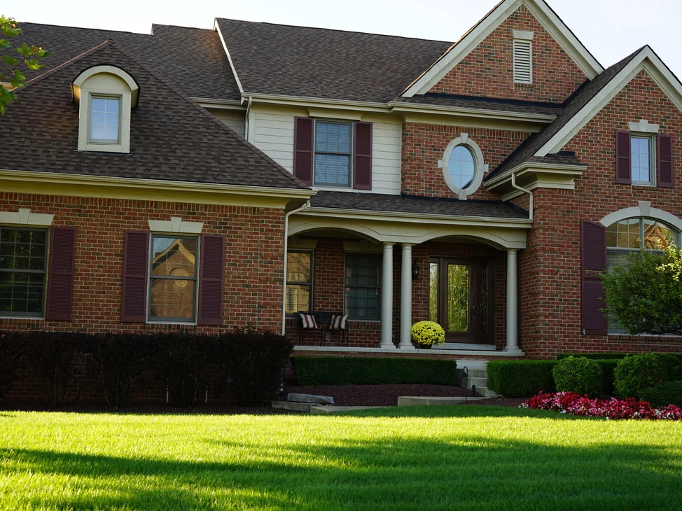 The front of the house in 2019 across the lawn, brick with cast stone lintels, an oval window over the columned porch, the wall reading as one red and brown range