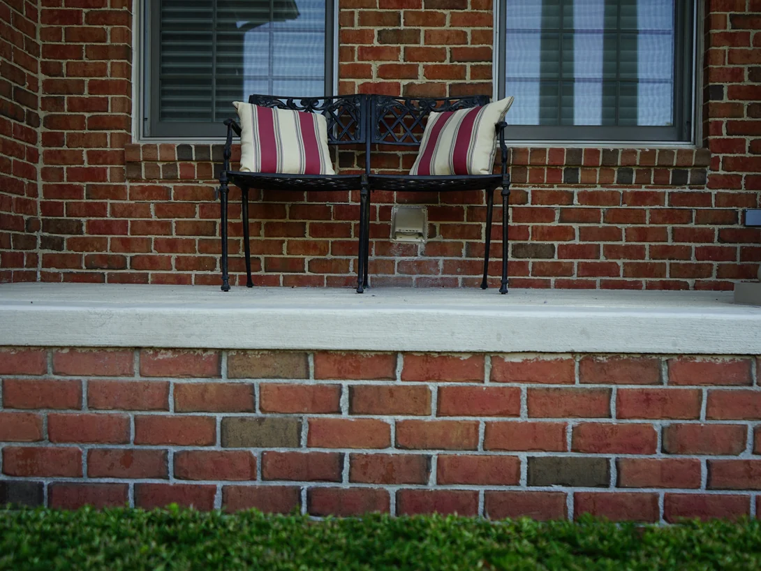 A metal bench on the front porch in 2019, the brick wall behind it and the sill course below the deck reading as one range of reds and browns