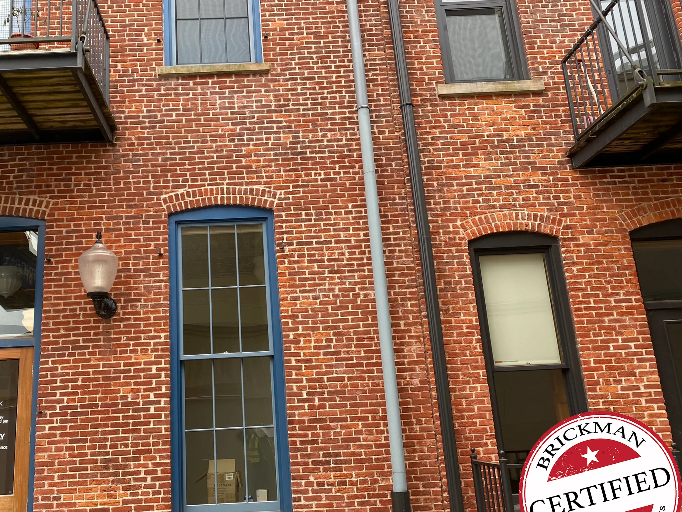 The front of the building in 2020, one range of red brick around segmental arched windows with blue frames, a downspout, iron balconies above and the certification stamp in the corner