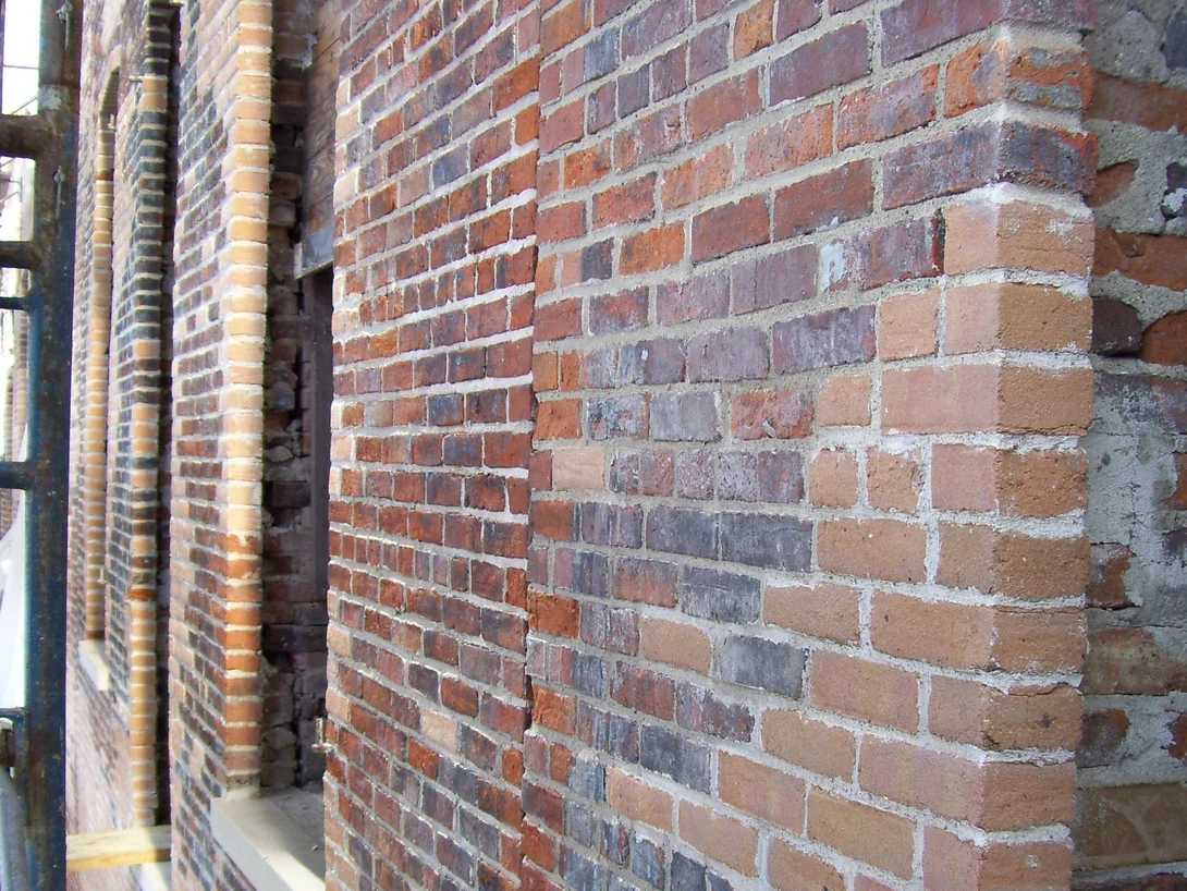 A brick pier and window reveal under scaffold in 2011, old red and purple brick beside a patch of new orange units and mismatched replacements at the corner