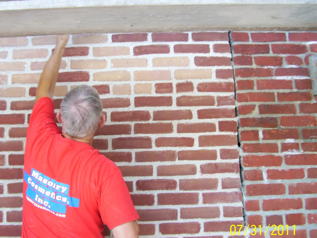 An applicator in a Masonry Cosmetics shirt working stain into a wall of mixed tan and red brick under a timber lintel in July 2011, a darker red wall beside it