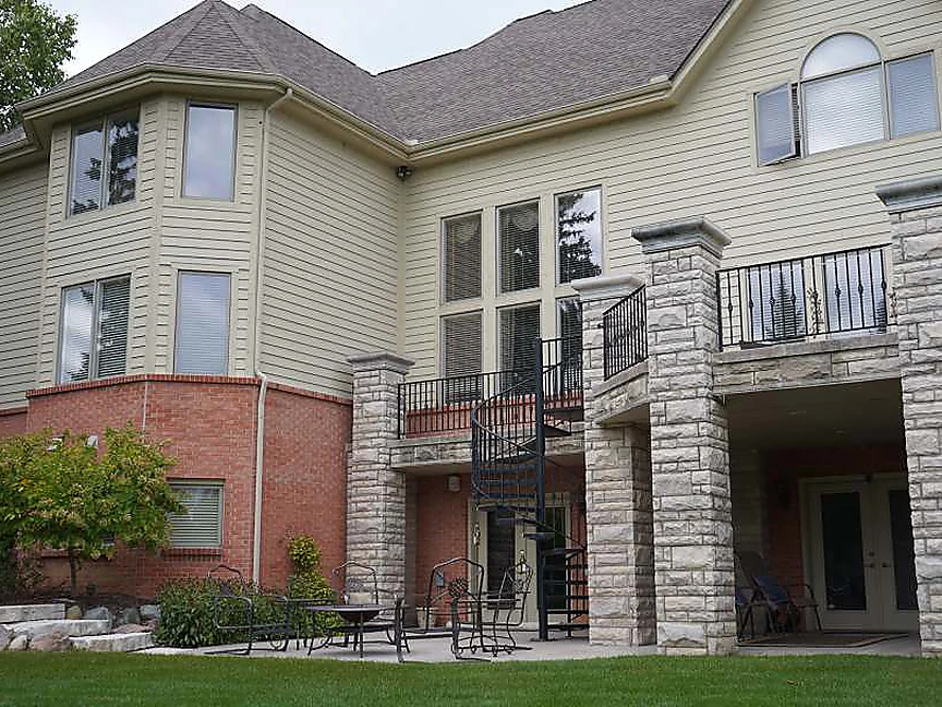 The back of the house in 2019, the same walkout brick reading as one even red under the siding, a mown lawn and planted beds where the rough grass was, stone-clad porch columns and a spiral stair added at the right