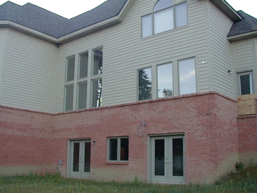 The walkout wall at the back of the house in 2002, pink brick under pale grey siding with washed-out units through the lower courses and rough unplanted grass along the foot