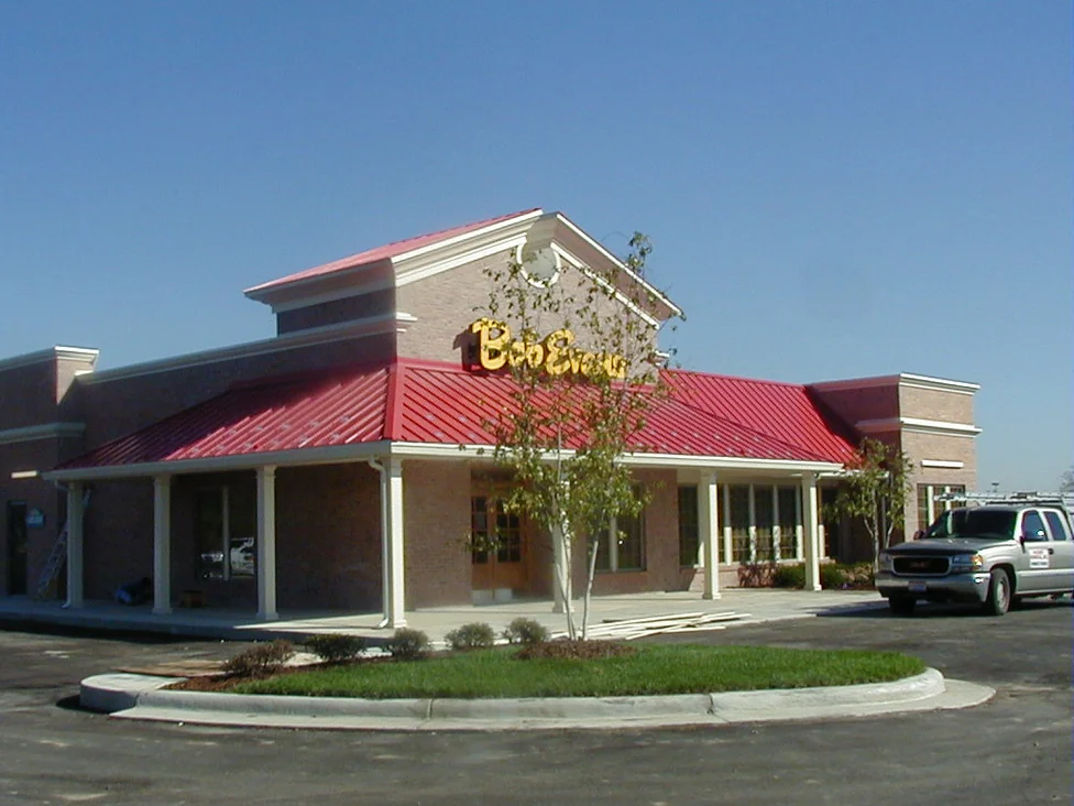 The finished restaurant from the parking lot in 2002, a low brick building with a red metal roof, a covered porch running along two sides and a gabled entrance