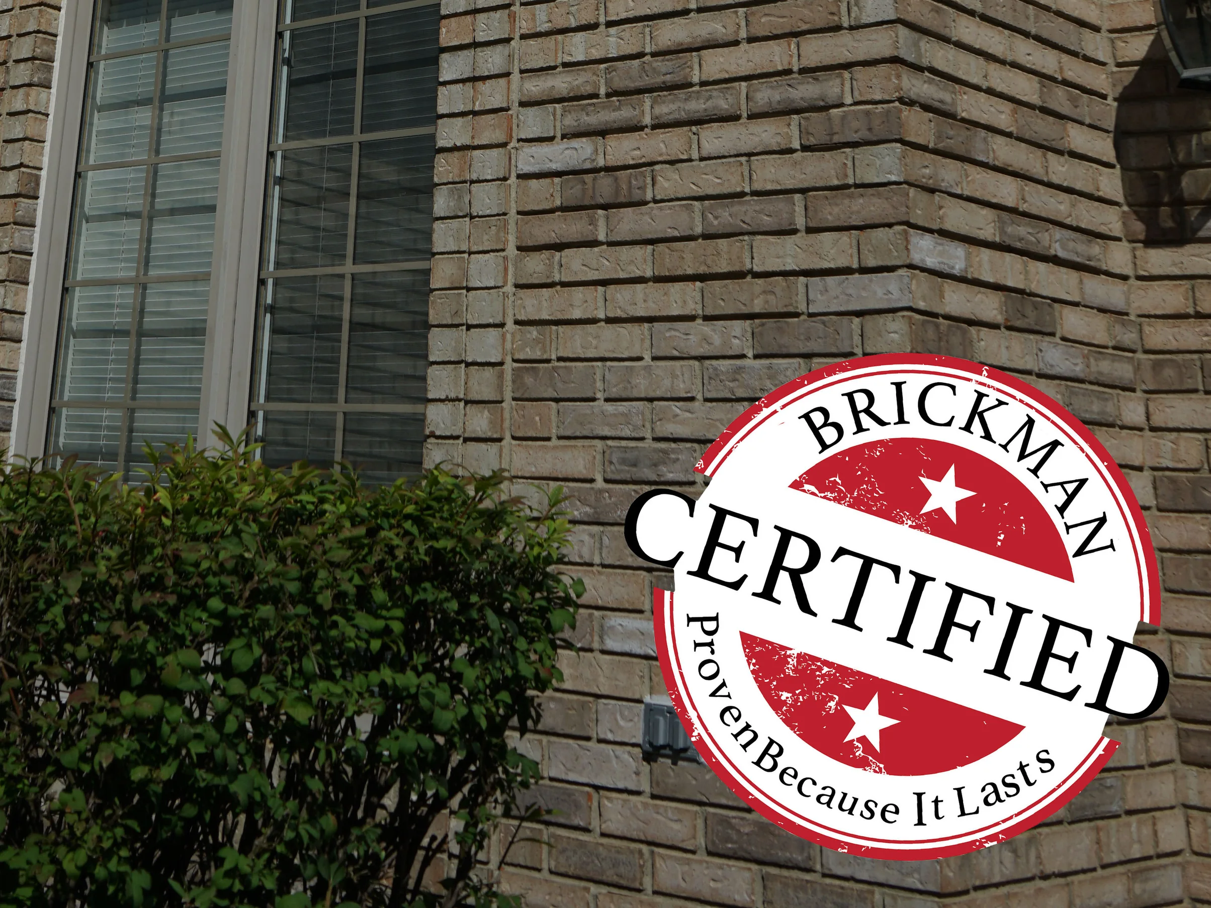 An outside brick corner of the house in 2019 beside a tall window, with a Brickman Certified stamp printed over the lower right of the frame