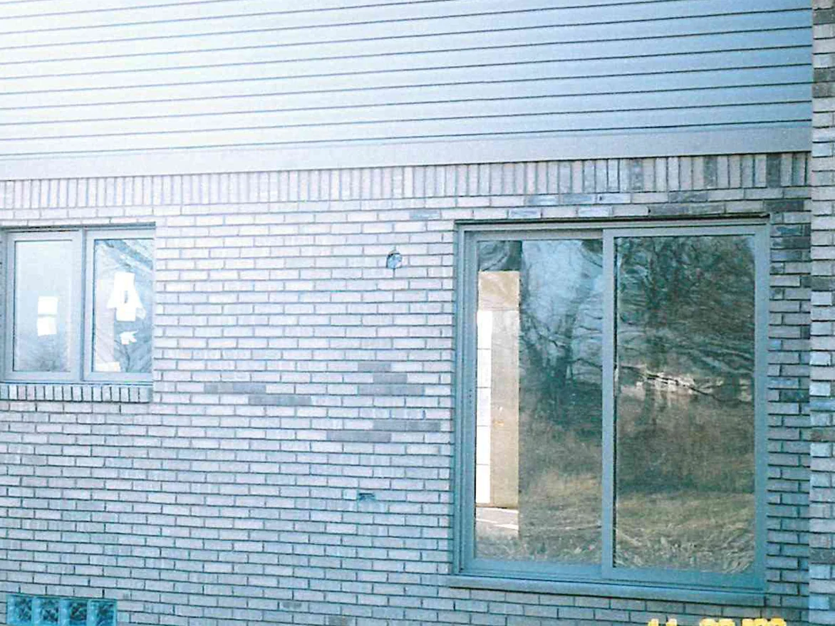 A side wall of the house in 2003, its clay brick a cold white buff below grey lap siding, with a sliding door and a glass block basement window