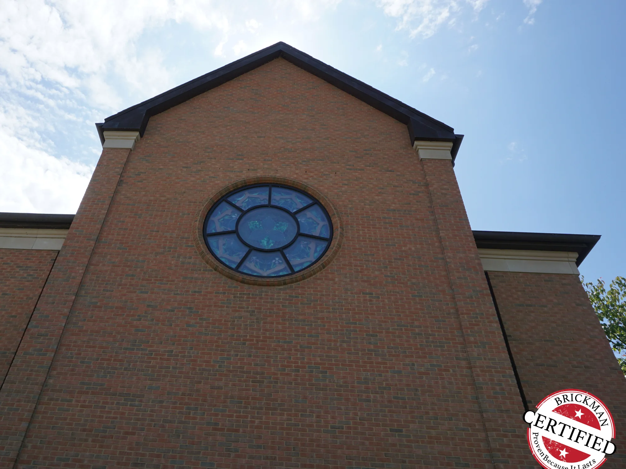 The gable end square on in 2019, the round stained glass window centred in brick that runs one consistent red and brown from the eaves to the ground, with the Brickman certification stamp in the corner