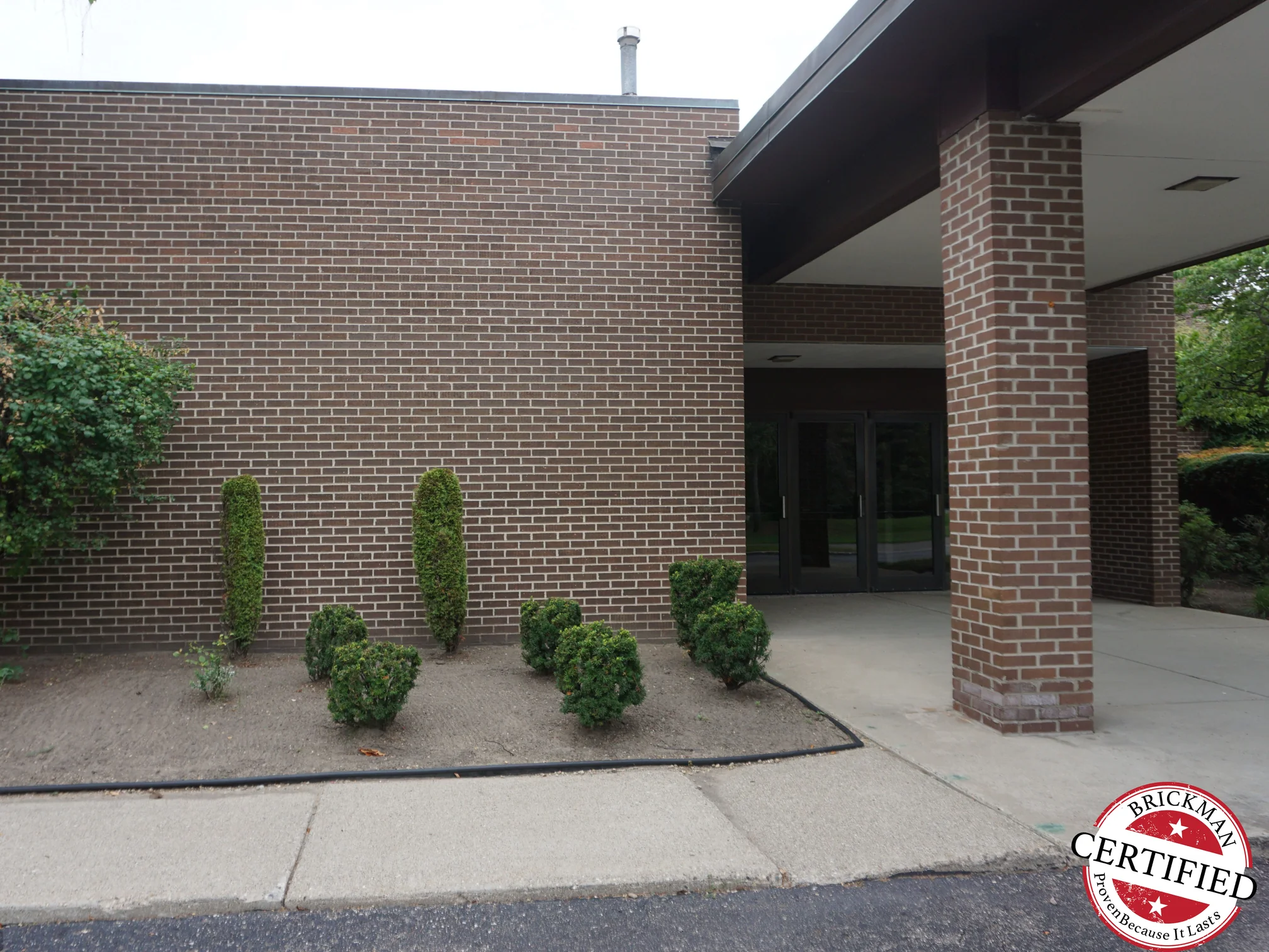 The church entrance in 2019, a canopy column standing against the long brick wall that runs to the glass doors, the Brickman certified stamp in the bottom corner of the frame
