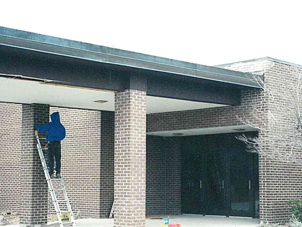 An applicator on a ladder working stain into a brick column under the entrance canopy, the columns reading lighter and redder than the wall behind them