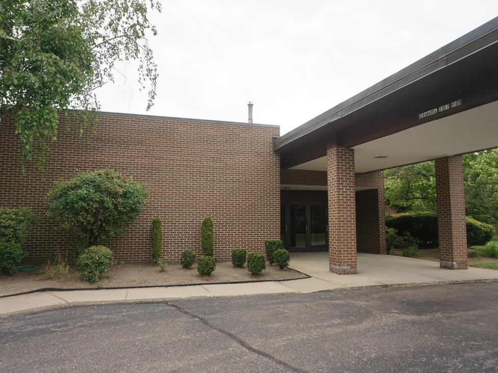 The side wall and entrance canopy from the drive in 2019, the wall and the canopy columns in one brown and red brick range under a dark fascia