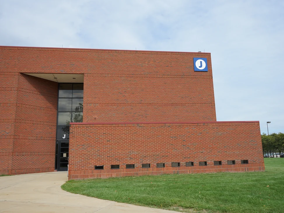 The end of the college building from the lawn in 2019, the recessed entrance, the low front wall and a blue roof sign lettered J, the brick one even red range.