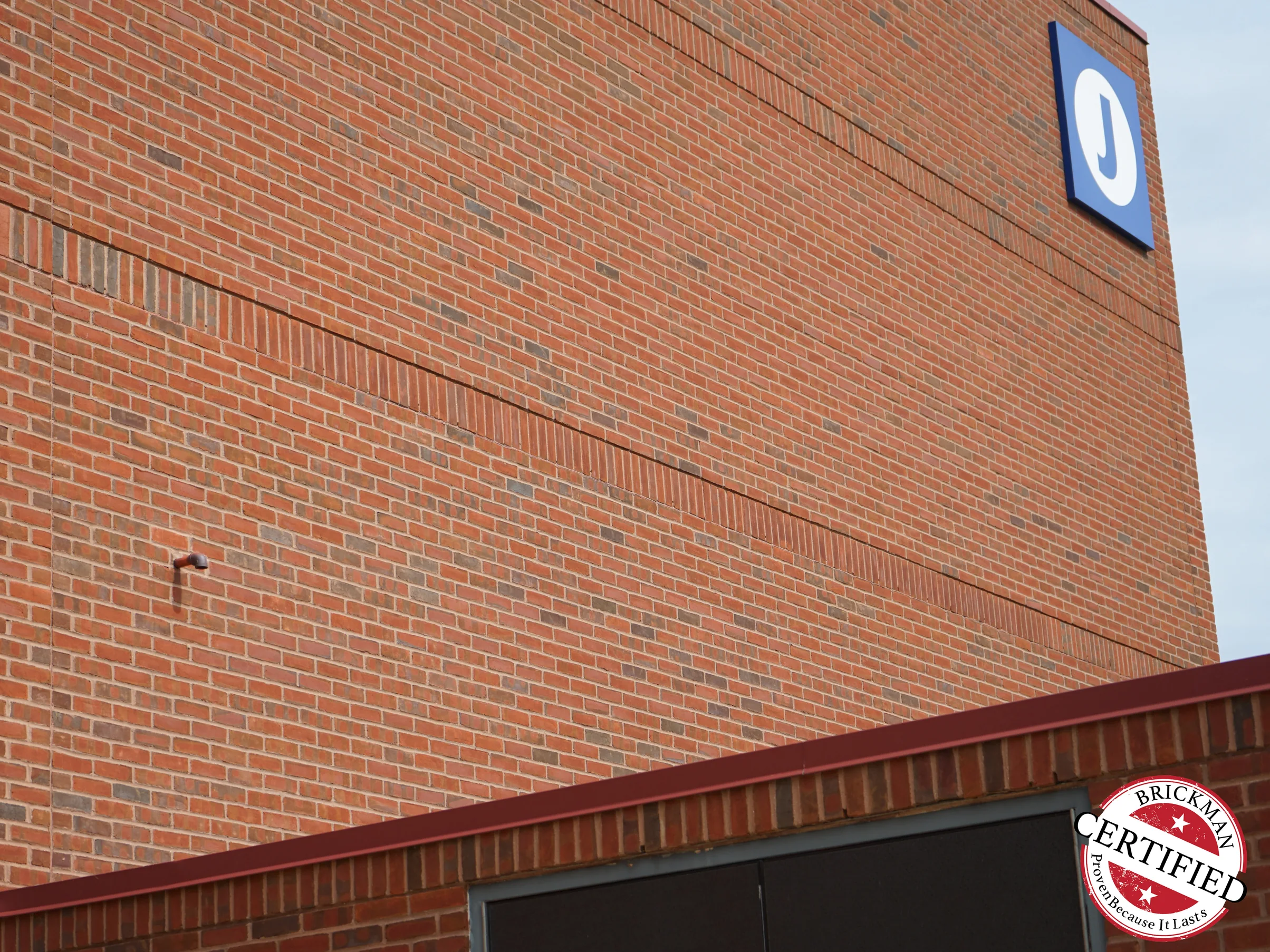 The upper wall and parapet in 2019 with the blue J sign at the corner, two soldier courses running the width of the wall in the same red range as the field brick