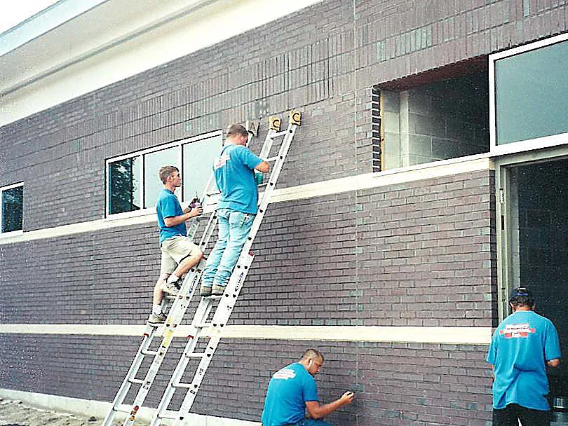 Four applicators in blue shirts working the joints of the rear elevation in 2000, two on extension ladders and one crouched at the base of the wall, with a cream accent band across the brick