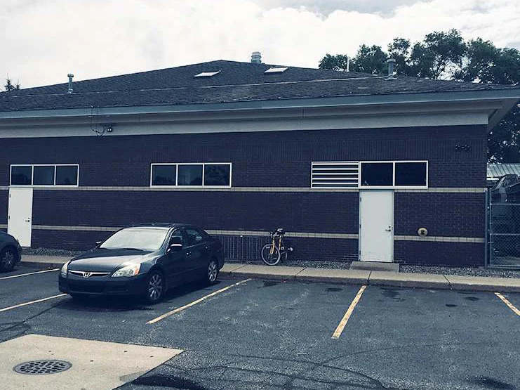 The rear elevation of the building in 2019 from the parking lot, dark brown brick with two cream accent bands, two white service doors, a louvered vent and a parked car in front