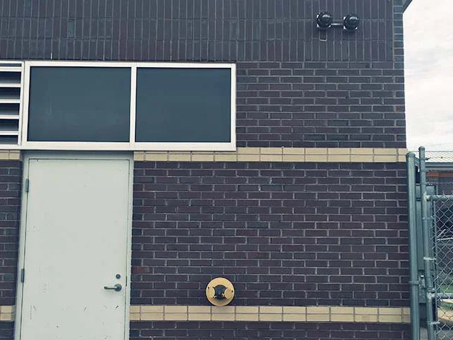 A pale service door in the rear wall in 2019, a cream accent band running above it and the sprinkler connection plate to its right, with a chain link fence at the corner