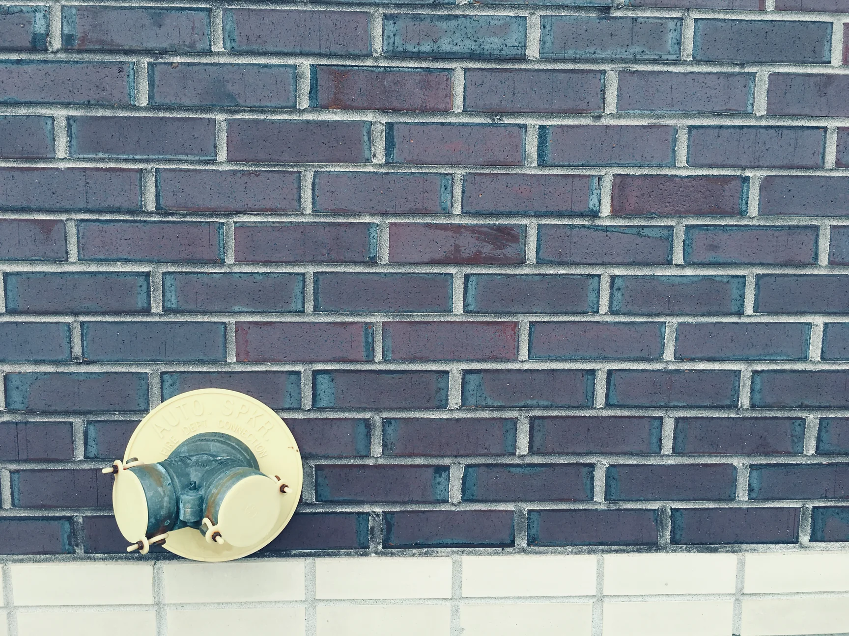 A fire department sprinkler connection on a cream plate set into the dark brown brick in 2019, the mortar joints reading grey inside the brick range, with a band of cream glazed brick below