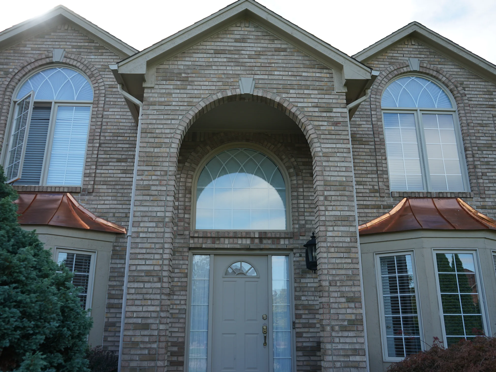 The front entry in 2019, a tall round-arched brick recess set between two gables, with copper roofs over the bay windows on either side