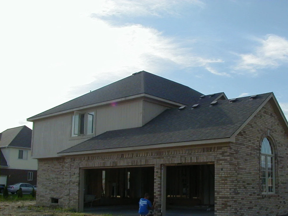 The house under construction in 2003, brick laid up to the open garage bays and the gable end while the upper storey is still bare sheathing, with an applicator kneeling in the garage opening