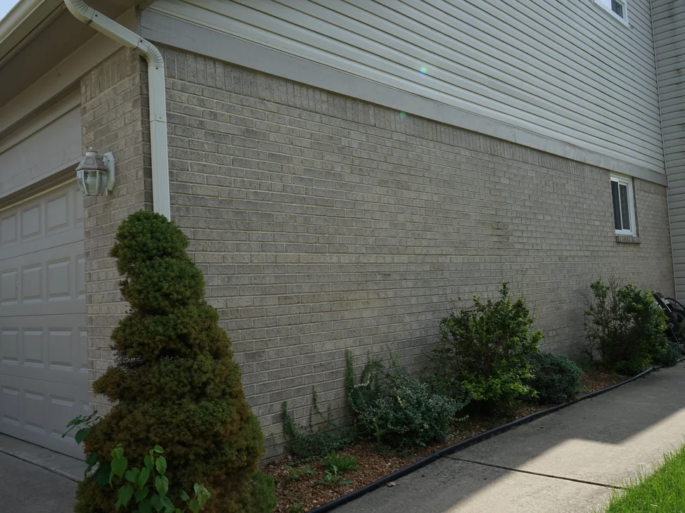 The side of the house from the driveway in 2019, grey brick running from the garage corner past a basement window to the far end of the wall.
