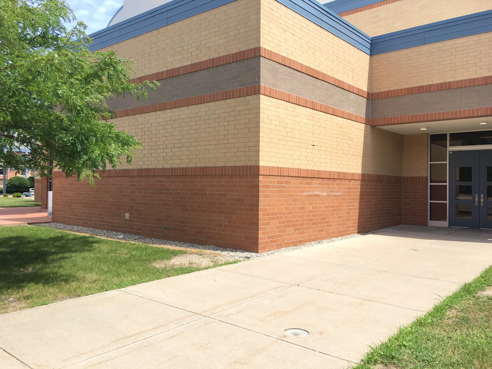 The outside corner of the high school in 2019, tan brick between a grey band and a red brown base, a tree at the left and the entrance doors at the right