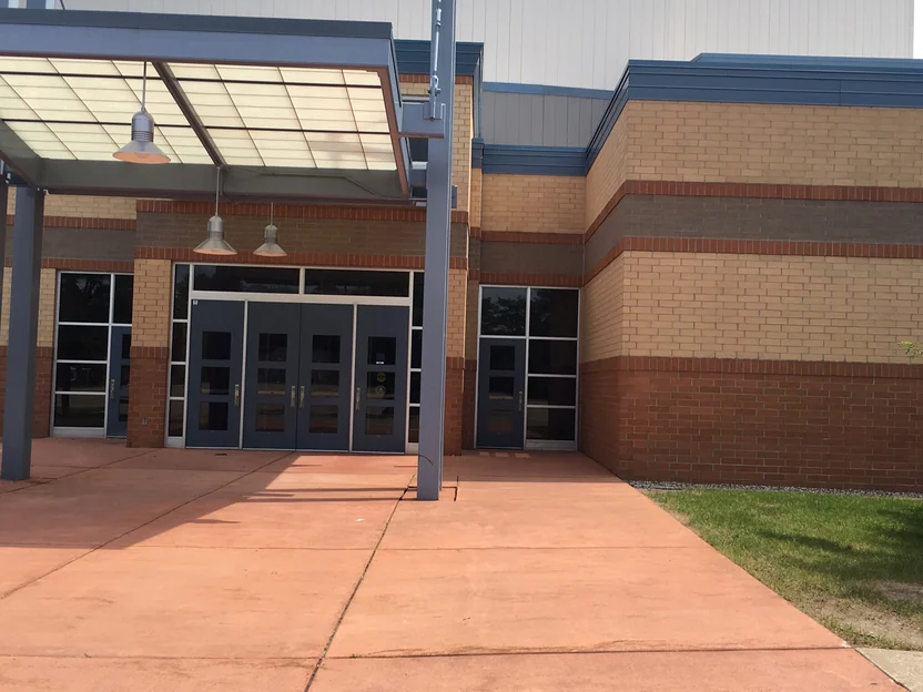 The main entrance in 2019, three sets of blue doors under a glazed canopy with pendant lights, the banded brick wall running off to the right