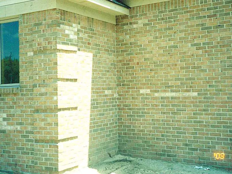 An outside corner of the house in May 2003 before staining, pale brick showing in the sunlit quoin column beside a window, a gas meter at the right and the camera date in the corner